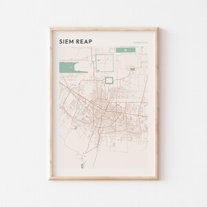 Pode incluir: Impressão emoldurada de um mapa de Siem Reap, Camboja. O mapa apresenta uma rede de estradas e ruas castanhas sobre um fundo bege claro. O texto "SIEM REAP" está no canto superior esquerdo, com "CAMBODIA" no canto superior direito. A moldura é de cor clara de madeira.