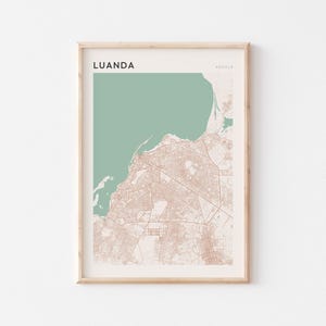 May include: Framed map art print of Luanda, Angola. The map features a muted color palette with a light beige background, a sea-green ocean, and detailed city streets in a reddish-brown hue. The word "LUANDA" is printed at the top.