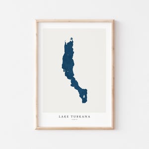 May include: A framed print showing a dark blue map of Lake Turkana in Kenya on a cream backdrop. The lake's form is detailed, with the text "LAKE TURKANA KENYA" beneath in a simple font. The frame is a light wood colour.