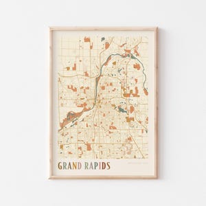 May include: A framed map print of Grand Rapids, Michigan, USA. The map has a beige background with brown and orange details, including roads, parks, and waterways. The words "GRAND RAPIDS" are printed in a gradient colour scheme.