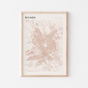 Op de afbeelding: Een ingelijste kaartprint van Riyad, Saoedi-Arabië, in een minimalistische stijl. De kaart toont een gedetailleerd stratennetwerk in bruintinten tegen een crèmekleurige achtergrond. Het woord "RIYADH" staat linksboven in het zwart gedrukt.