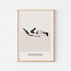 May include: A framed art print with a minimalist black map of Indonesia on a beige background. The word "INDONESIA" is printed below the map in a simple font. The print is in a light wood frame, suitable for home decor.