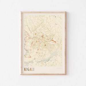 May include: A framed map print of Kigali, Rwanda, in a neutral colour palette. The map features detailed topographical lines and water features, with the city name in a gold font at the bottom. The print is encased in a light wood frame.