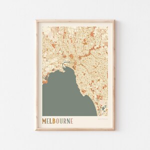 May include: A framed map print of Melbourne, Australia, in muted tones of beige, orange, and green. The city's layout is detailed, with the word "MELBOURNE" at the bottom in a multi-colored font. The print is in a light wooden frame.