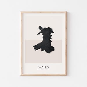 May include: A framed print showing a black silhouette of Wales set against a neutral backdrop. The word "WALES" is printed in a simple font at the bottom. The artwork is encased in a light-coloured wooden frame.
