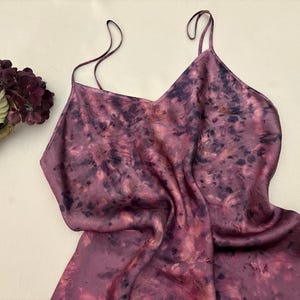 May include: A sleeveless slip dress featuring a mottled tie-dye pattern in shades of deep purple, magenta, and dark violet. The garment has a V-neckline and thin spaghetti straps, accompanied by a single dark purple hydrangea flower stem.