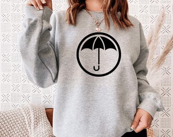 Könnte beinhalten: Ein hellgraues Sweatshirt mit einem schwarzen Regenschirm-Logo in einem schwarzen Kreis. Das langärmlige Oberteil hat einen Rundhalsausschnitt und gerippte Bündchen. Das Regenschirm-Logo ist mittig auf der Brust platziert.