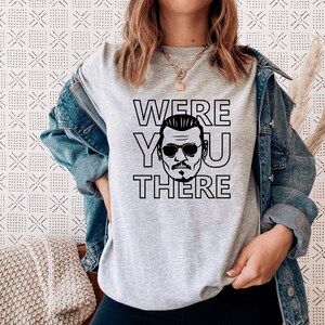 Op de afbeelding: Een lichtgrijs t-shirt met korte mouwen en een zwarte grafische print van een man met een zonnebril, met de tekst "WERE YOU THERE" rondom de illustratie. Het t-shirt wordt gedragen onder een blauw spijkerjack met lange mouwen en knopen.