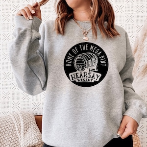 Op de afbeelding: Een lichtgrijs sweatshirt met ronde hals en een zwarte cirkelvormige afbeelding op de borst. De afbeelding toont een houten vat met de tekst "HOME OF THE MEGA PINT" in een boog erboven en "HEARSAY WINERY" op een banner onderaan.
