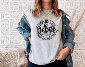 May include: A light grey t-shirt featuring a black circular graphic with the text "Happy Hour 24/7 Depps Whiskey & Wine Home of the MEGA PINT". The shirt is being worn.