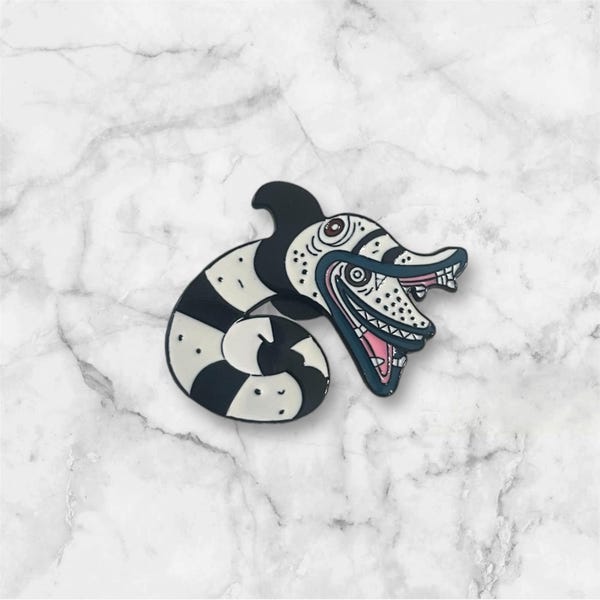 May include: Black and white enamel pin featuring a cartoon character with a long, thin body and a large, toothy grin. The character has a black and white striped body and a blue fin on its head.