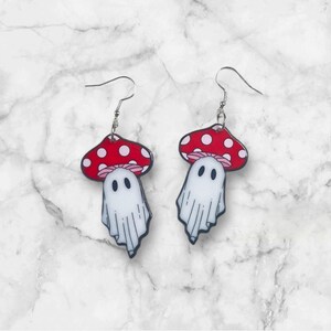 May include: A pair of dangle earrings featuring a red and white mushroom with white ghost face. The mushroom has white polka dots.