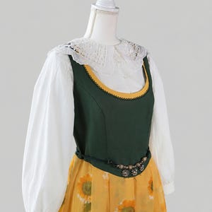 May include: A dress featuring a white long-sleeved blouse with a lace collar, a dark green bodice with yellow trim, and a yellow skirt with a sunflower print. A thin black belt with metallic beads and flower charms sits at the waist.