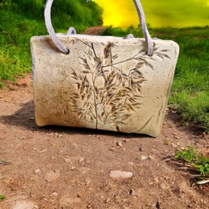Könnte beinhalten: Beige Keramik-Tragetasche mit Bambusblatt-Design. Die Tasche hat eine rechteckige Form mit abgerundeten Ecken und grauen Seilgriffen. Sie steht auf einem Feldweg mit grünem Gras im Hintergrund.