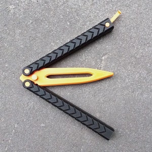 Puede incluir: Un cuchillo mariposa negro y dorado con un mango texturizado y una hoja amarilla. El cuchillo est&aacute; abierto y la hoja est&aacute; extendida.