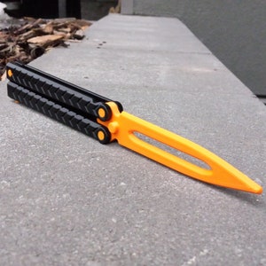 Puede incluir: Un cuchillo mariposa negro y naranja con un mango texturizado. El cuchillo est&aacute; cerrado y se encuentra sobre una superficie de hormig&oacute;n gris.