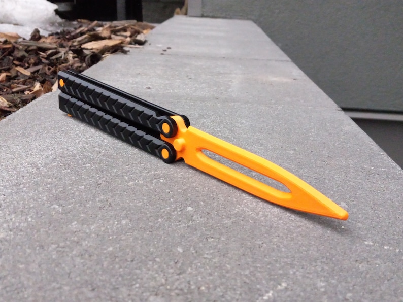 Puede incluir: Un cuchillo mariposa negro y naranja con un mango texturizado. El cuchillo est&aacute; cerrado y se encuentra sobre una superficie de hormig&oacute;n gris.