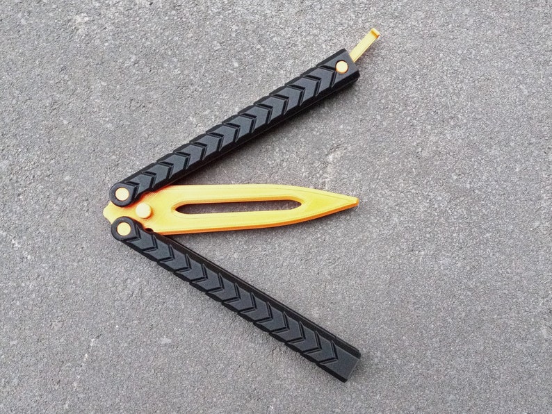 Puede incluir: Un cuchillo mariposa negro y dorado con un mango texturizado y una hoja amarilla. El cuchillo est&aacute; abierto y la hoja est&aacute; extendida.