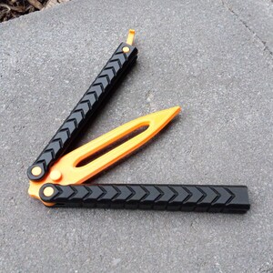 Puede incluir: Un cuchillo mariposa negro y naranja con un mango texturizado. El cuchillo est&aacute; abierto y la hoja es visible.