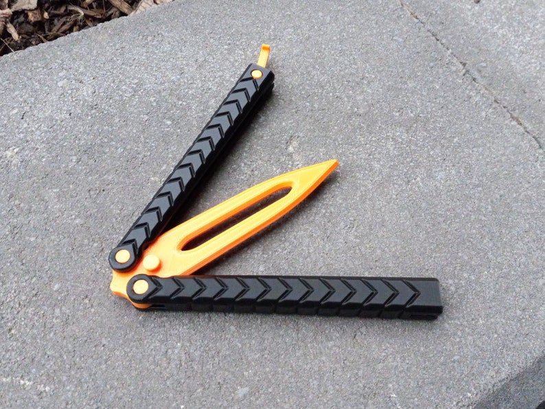 Puede incluir: Un cuchillo mariposa negro y naranja con un mango texturizado. El cuchillo est&aacute; abierto y la hoja es visible.