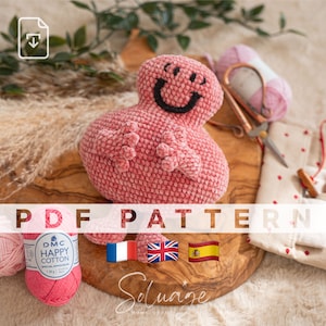 May include: A pink crocheted amigurumi toy in the shape of a smiling character with black eyes and a black mouth. The toy is sitting on a wooden surface with a ball of pink yarn and a pair of scissors. The text "PDF PATTERN" is written in white letters on a black background.