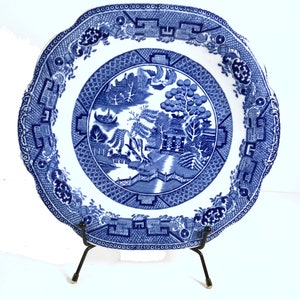 May include: A blue and white porcelain plate with a willow pattern design. The plate has a scalloped edge and is on a black metal stand.