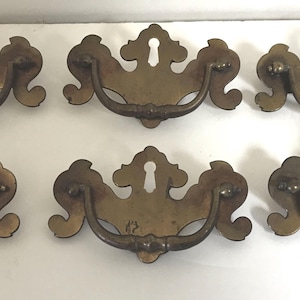 May include: Six antique brass drawer pulls with a keyhole design. Each pull has a curved handle and decorative backplate. The brass has a warm, aged patina, suggesting vintage or antique hardware. Ideal for restoring furniture.