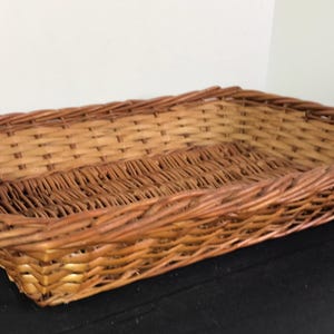 May include: A rectangular woven wicker basket with a natural brown color. The basket has a sturdy construction with a woven base and sides, perfect for storage or display. The basket's dimensions are approximately 16 inches long, 10 inches wide, and 4 inches deep.