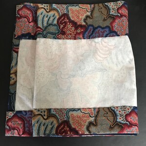 May include: A folded textile with a white center and patterned borders. The borders feature a mix of red, blue, and tan floral and abstract designs. The white center has a subtle cloud-like pattern.