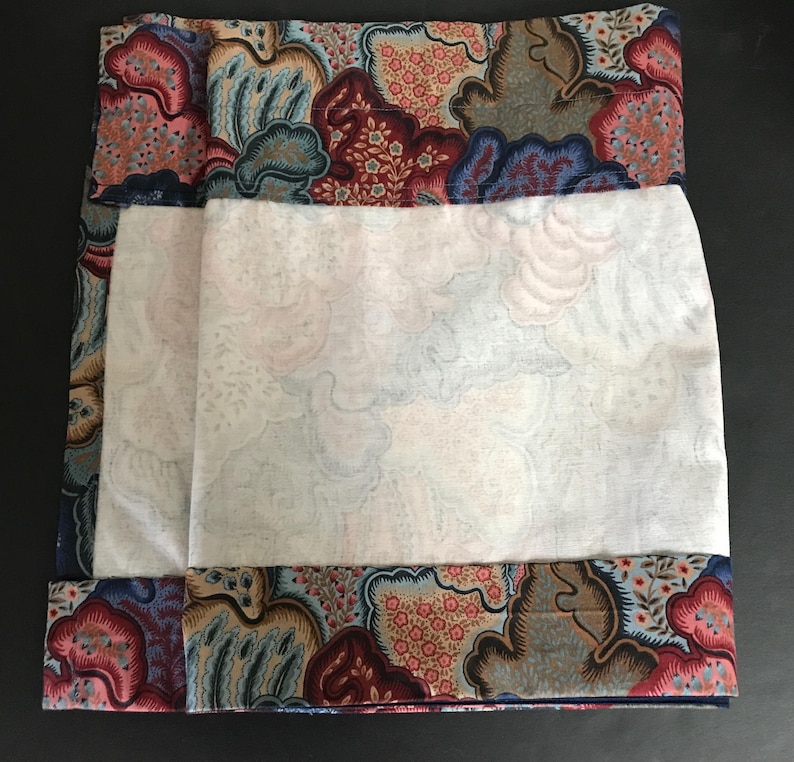 May include: A folded textile with a white center and patterned borders. The borders feature a mix of red, blue, and tan floral and abstract designs. The white center has a subtle cloud-like pattern.