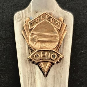 May include: A silver spoon with a gold-colored emblem featuring the words "TOLEDO OHIO" and a depiction of a bridge.