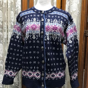 May include: A navy blue cardigan sweater with a detailed geometric pattern in white, light blue, and purple. The sweater has long sleeves, a round neckline, and is fastened with silver-toned buttons. The design is a classic Nordic style.