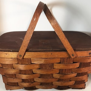 May include: A rectangular picnic basket with a wooden lid and handle. The basket is woven with a light brown material, and the lid and handle are a darker brown. The handle is attached to the basket with metal rivets.