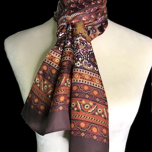 May include: A patterned silk scarf in shades of brown, orange, and purple, draped on a mannequin. The scarf features intricate floral and geometric designs. The scarf is approximately 60 inches long.