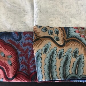 May include: Close-up of fabric samples with intricate floral and paisley patterns in shades of red, blue, tan, and pink. The fabric has a vintage aesthetic, with a detailed design and a soft texture.