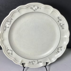 May include: A round, off-white ceramic dinner plate with a scalloped edge and a speckled finish. The rim features five small, evenly spaced floral motifs in grey, consisting of tiny white flowers and leaves arranged in a delicate, repeating pattern.