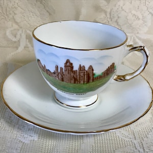 May include: A white teacup and saucer with a gold rim. The teacup features a painted image of Arbroath Abbey in Scotland.