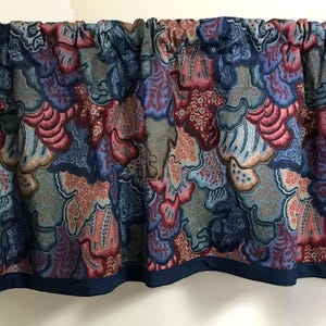 May include: A valance with a repeating pattern of abstract shapes in shades of blue, red, and gray. The fabric is gathered at the top and has a dark blue border along the bottom edge. This decorative valance is designed for a window.