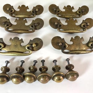 May include: Set of six antique brass drawer pulls and six matching knobs. The drawer pulls have a decorative, ornate design with a curved handle. The knobs are round with a decorative base. Perfect for furniture restoration projects.