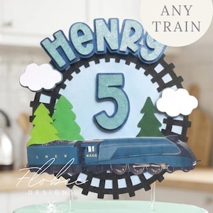 May include: A cake topper featuring a blue train, the name "Henry", and the number "5". The design includes trees, clouds, and the text "Pick Any Train". The topper is set on a light green cake, with a black cogwheel design.