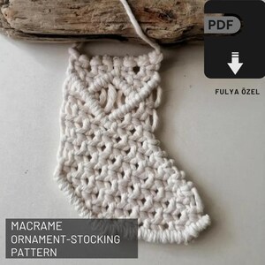 Pode incluir: Um padrão de ornamento de meia de Natal em macramé branco. O padrão é para um ornamento de meia que é feito com nós de macramé. O padrão está disponível para download em PDF.