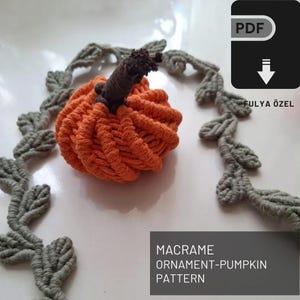 Pode incluir: Um padrão de ornamento de abóbora de macramé para download em PDF. O padrão inclui instruções para criar uma pequena abóbora laranja com um caule marrom. O padrão é cercado por uma borda de folhas de macramé cinzas.