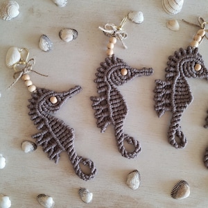 May include: Three brown macrame seahorse keychains with wooden beads hanging from gold key rings. The keychains are arranged on a white wooden surface with seashells scattered around them.