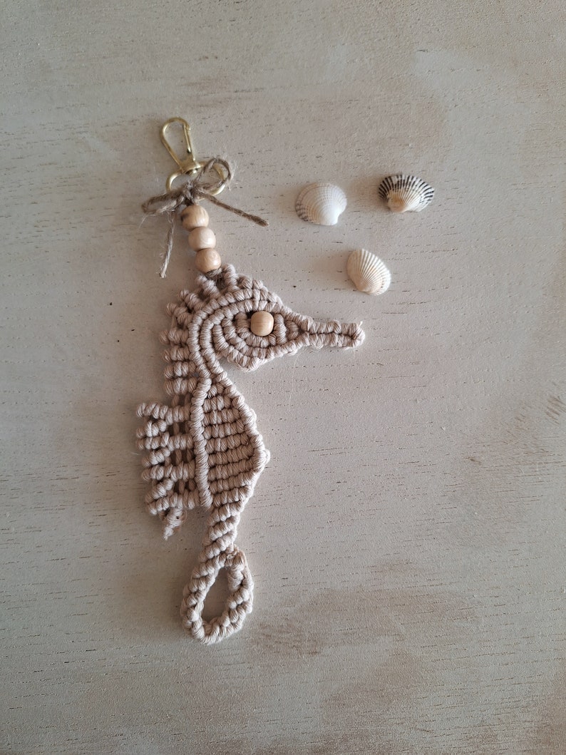 May include: A beige macrame seahorse keychain with wooden beads and a gold metal ring. The keychain is on a light wood surface with three small white seashells.