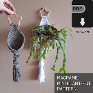 May include: Two macrame plant hangers with wooden star tops. One is white with a white tassel and the other is green with a green tassel. The white hanger has a small plant in it. The text "MACRAME MINI PLANT-POT PATTERN" is at the bottom of the image. The text "PDF" is in a black box with a download arrow in the top right corner. The text "FULYA ÖZEL" is below the box.
