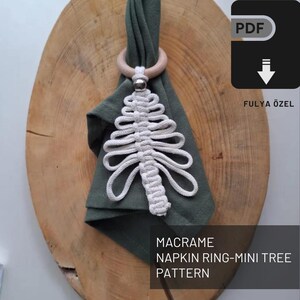 Könnte beinhalten: Ein weißer Makramee-Serviettenring in Form eines kleinen Baumes, der an einem Holzring an einer grünen Serviette hängt. Die Serviette liegt auf einer Holzoberfläche. Der Text "MACRAME NAPKIN RING-MINI TREE PATTERN" befindet sich unter dem Bild.