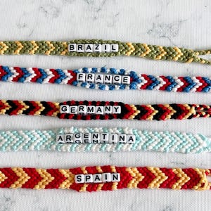 K&ouml;nnte beinhalten: Handgefertigte Freundschaftsarmb&auml;nder in verschiedenen Farben, jeweils mit einem L&auml;ndernamen, der in wei&szlig;en Buchstabenperlen geschrieben ist. Die Armb&auml;nder umfassen Brasilien, Frankreich, Deutschland, Argentinien und Spanien. Chevron-Muster.