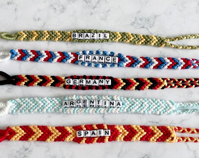 K&ouml;nnte beinhalten: Handgefertigte Freundschaftsarmb&auml;nder in verschiedenen Farben, jeweils mit einem L&auml;ndernamen, der in wei&szlig;en Buchstabenperlen geschrieben ist. Die Armb&auml;nder umfassen Brasilien, Frankreich, Deutschland, Argentinien und Spanien. Chevron-Muster.