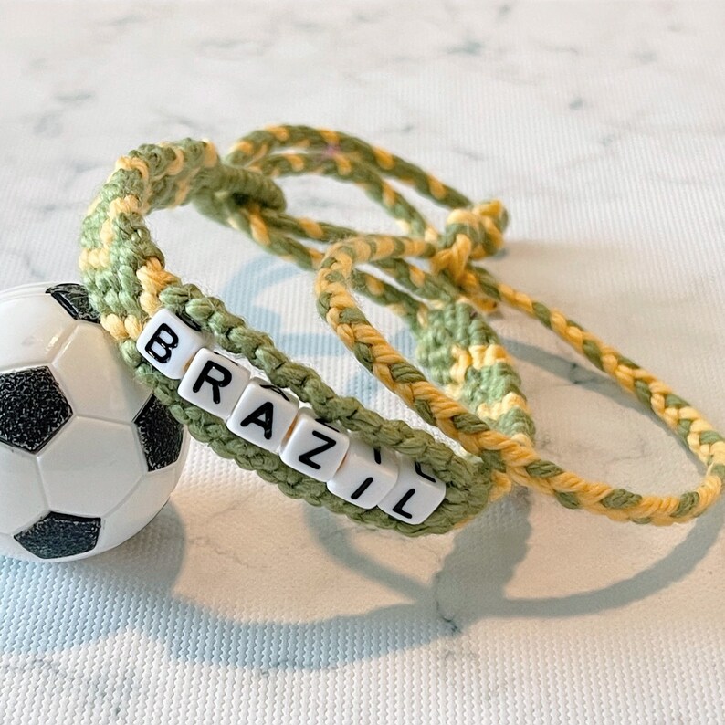 K&ouml;nnte beinhalten: Ein handgefertigtes Armband mit dem Wort "BRAZIL" in wei&szlig;en Buchstabenperlen. Das Armband ist gr&uuml;n und gelb, daneben liegt ein kleiner Fu&szlig;ball.