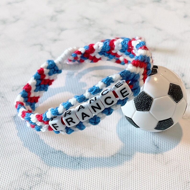 K&ouml;nnte beinhalten: Ein geflochtenes Armband in den Farben der franz&ouml;sischen Flagge: Rot, Wei&szlig; und Blau. Das Wort "FRANCE" ist mit wei&szlig;en Buchstabenperlen geschrieben. Ein kleiner Fu&szlig;ball-Charm ist am Armband befestigt.